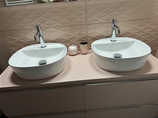 Twee witte opbouwspoelbakken op een beige aanrechtblad met chromen kranen en kleine zeepdispensers.