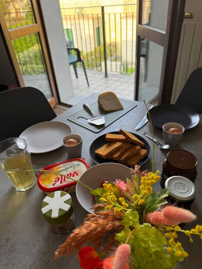 Een eettafel gedekt buiten, met brood, yoghurt, drankjes en een bloemstuk in het midden, bij een balkondeur.