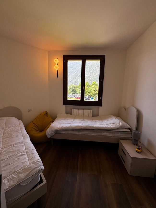 Kleine slaapkamer met twee bedden, een raam, een donkere houten vloer en een warme wandlamp.
