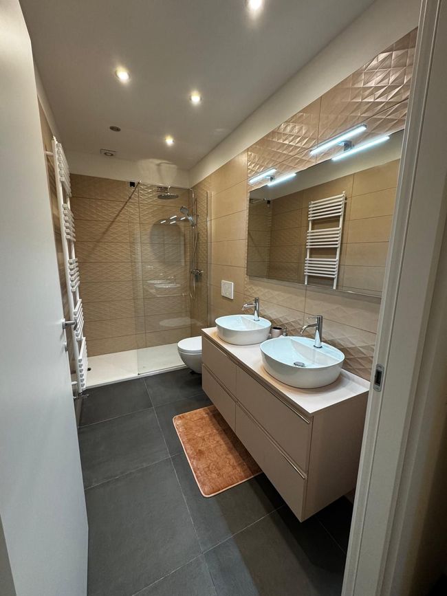 Moderne badkamer met dubbele wastafel, grote spiegel, beige tegels en zwarte vloertegels.