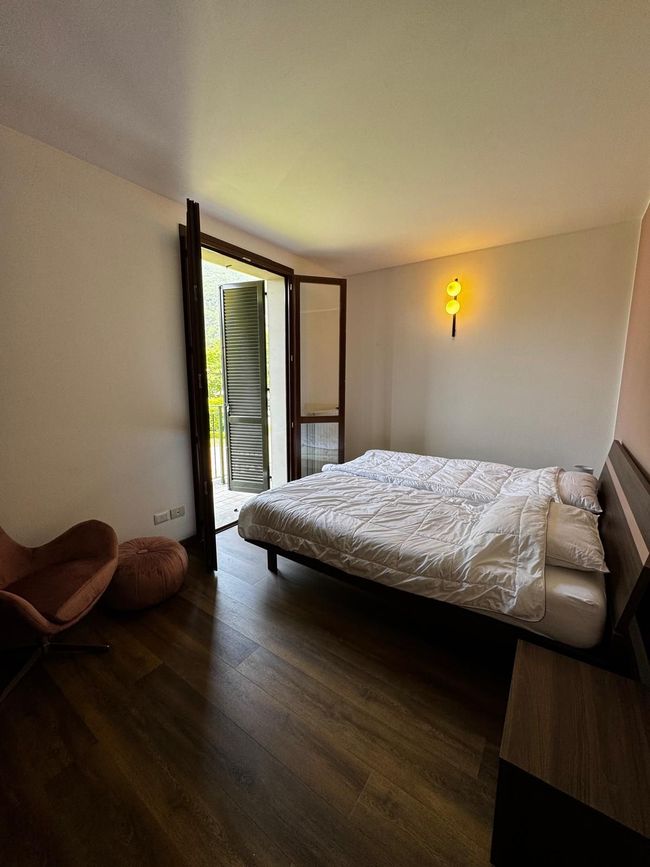 Minimalistische slaapkamer met bed, openslaande balkondeur, donkere houten vloer en wandlamp.