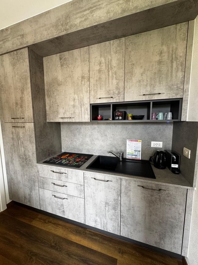 Moderne, ingebouwde keukennis met witte kasten, zwart aanrechtblad, spoelbak en kookplaat tegen een grijze muur.
