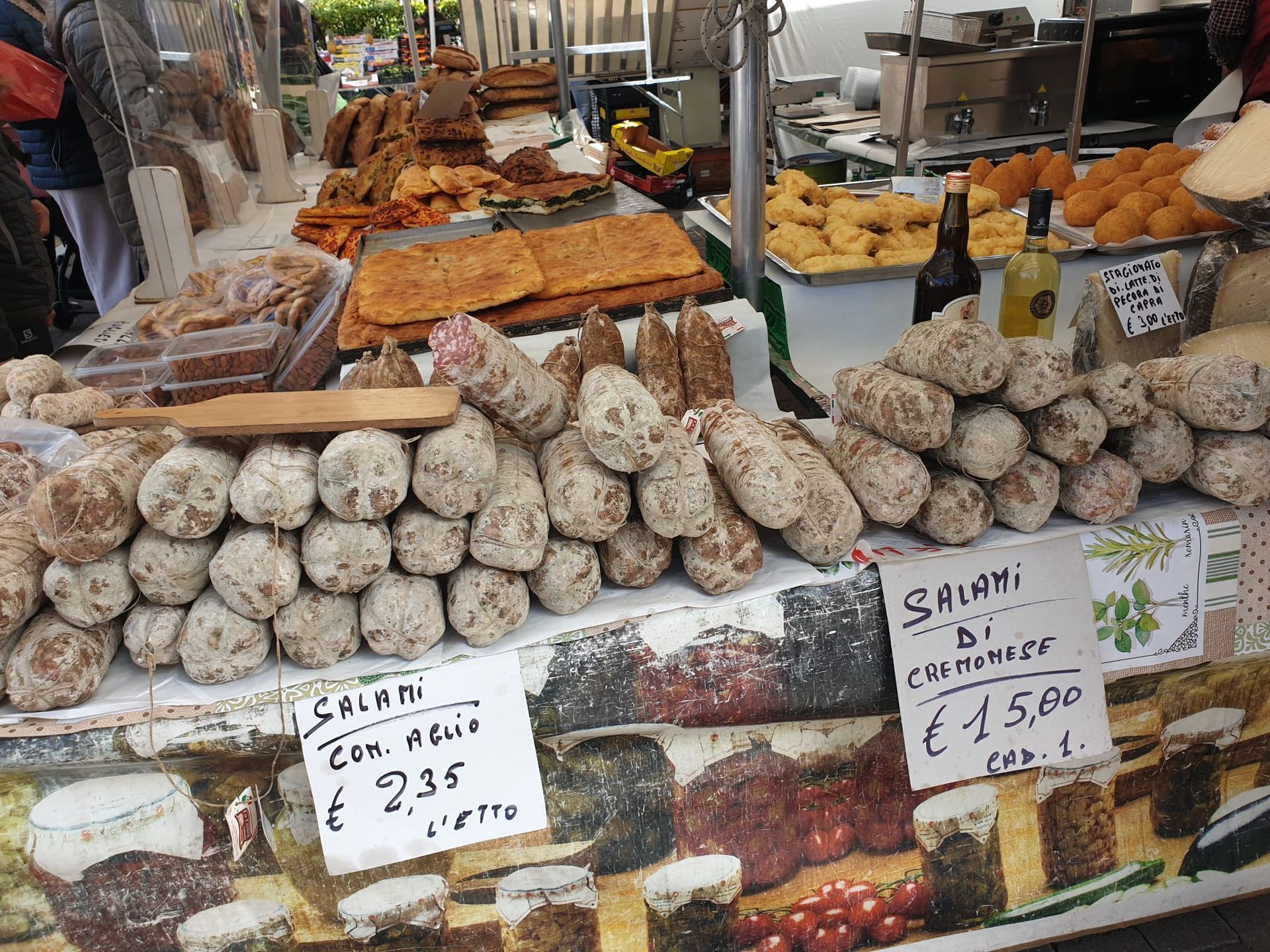 Marktkraam met diverse kazen, salami en verpakte voedingsmiddelen uitgestald, met handgeschreven prijskaartjes.