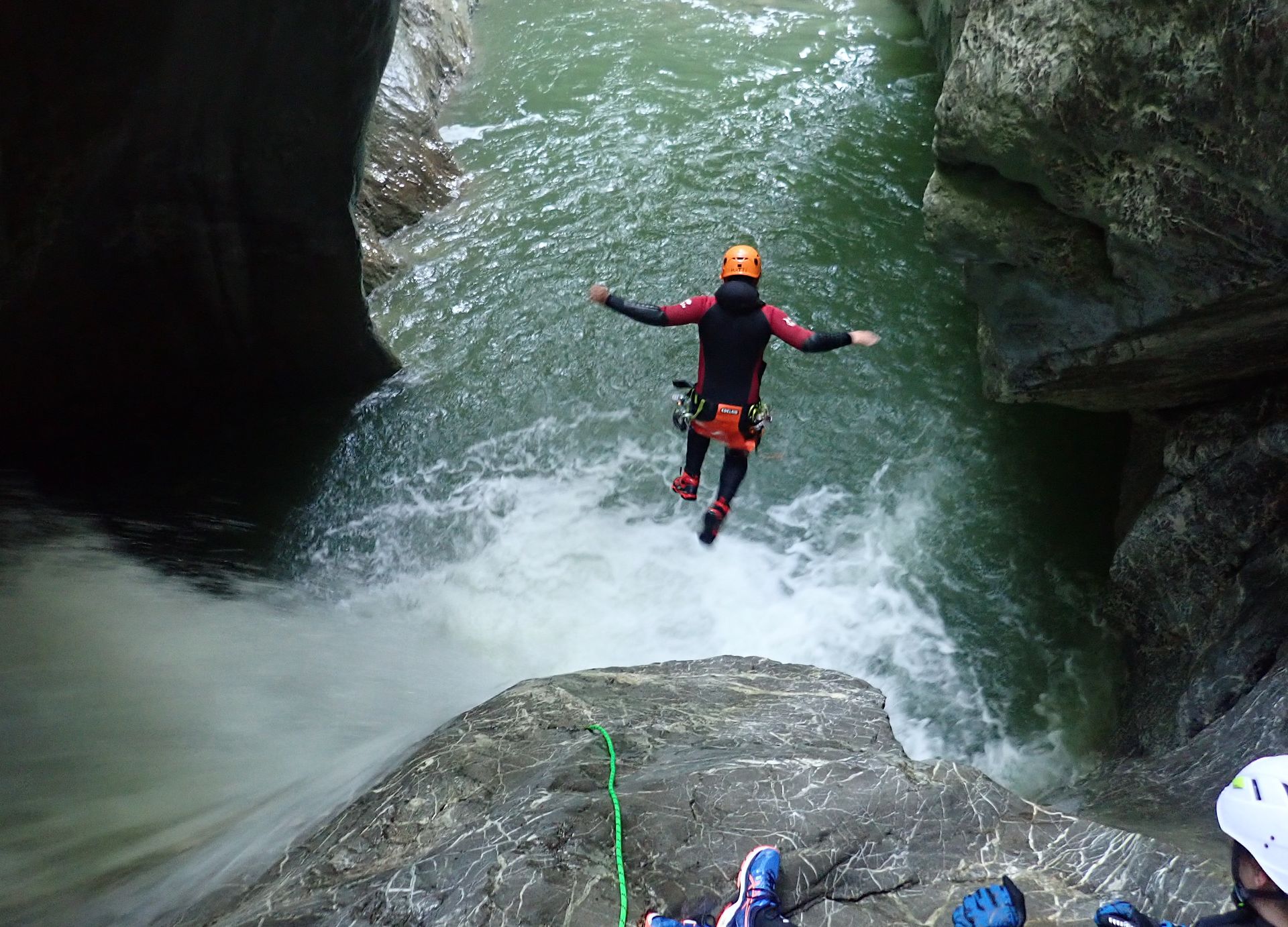 Canyoningbeoefenaar met oranje helm daalt af in een smalle rivierkloof met kolkend water.