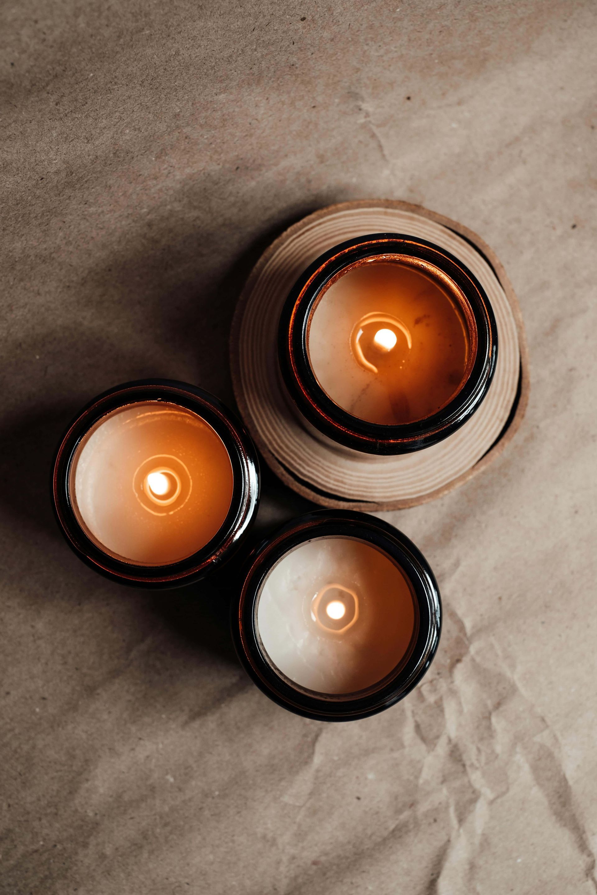 Three lit candles in decorative containers on a crinkled brown surface, viewed from above.