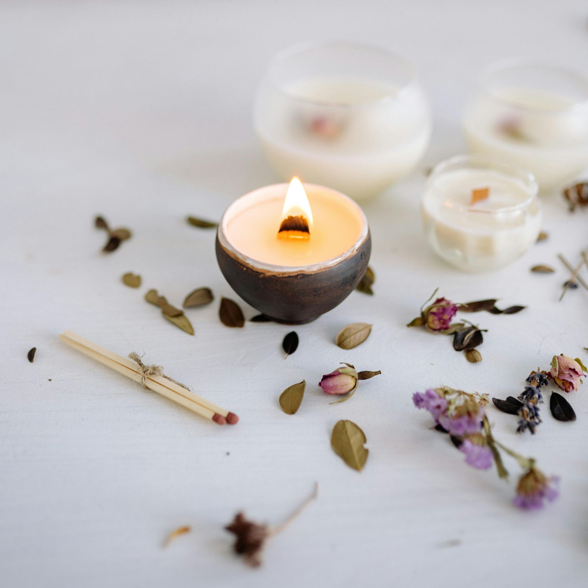 A lit candle with a wooden wick, surrounded by other candles, dried flowers, and a match on a white surface.