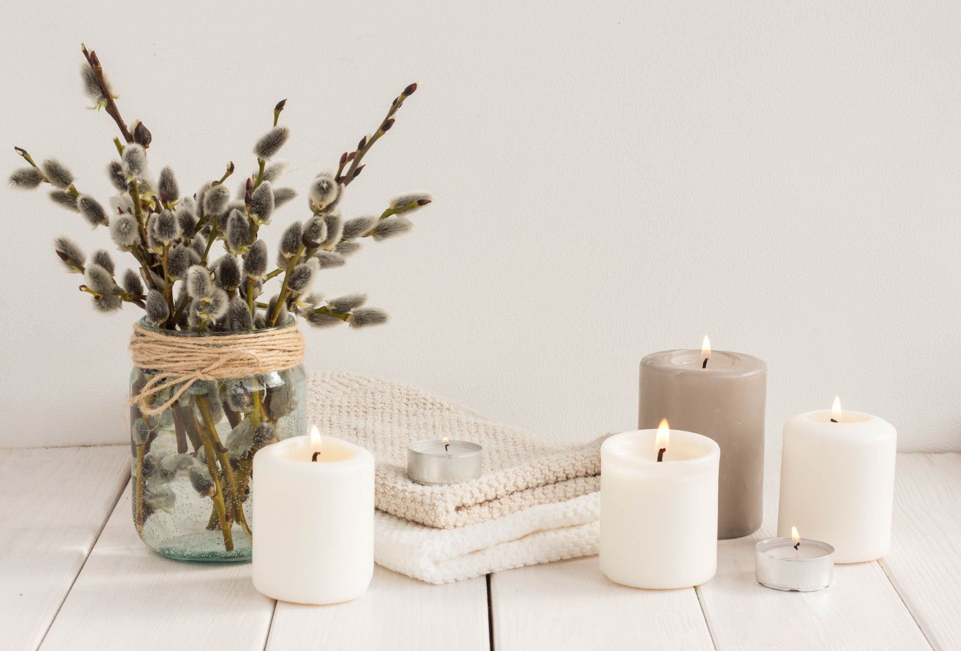White candles of varying sizes and a vase of pussy willow branches sit on a white wooden table.