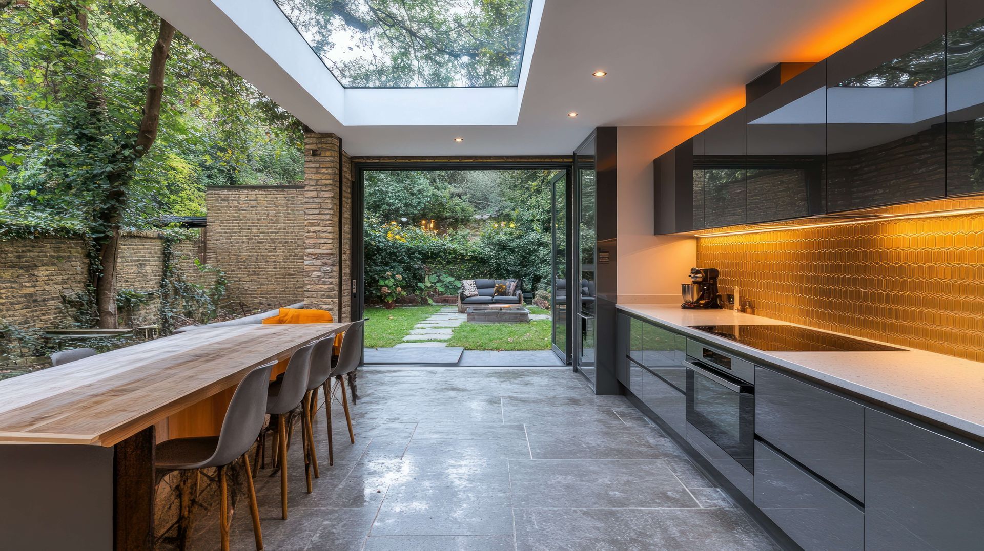 Modern kitchen with long wooden dining table, sleek cabinets, and open view to a green backyard.