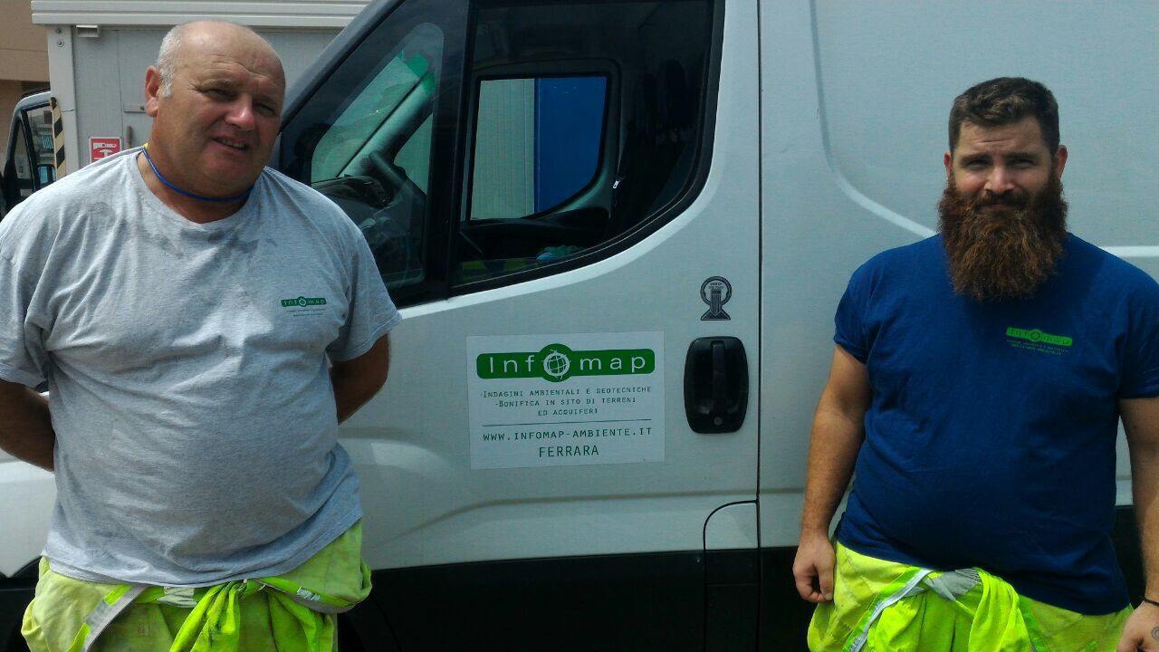 Due operai con pantaloni fluorescenti sono in piedi accanto a un camion bianco, uno calvo con una camicia grigia, l'altro barbuto con una camicia blu.