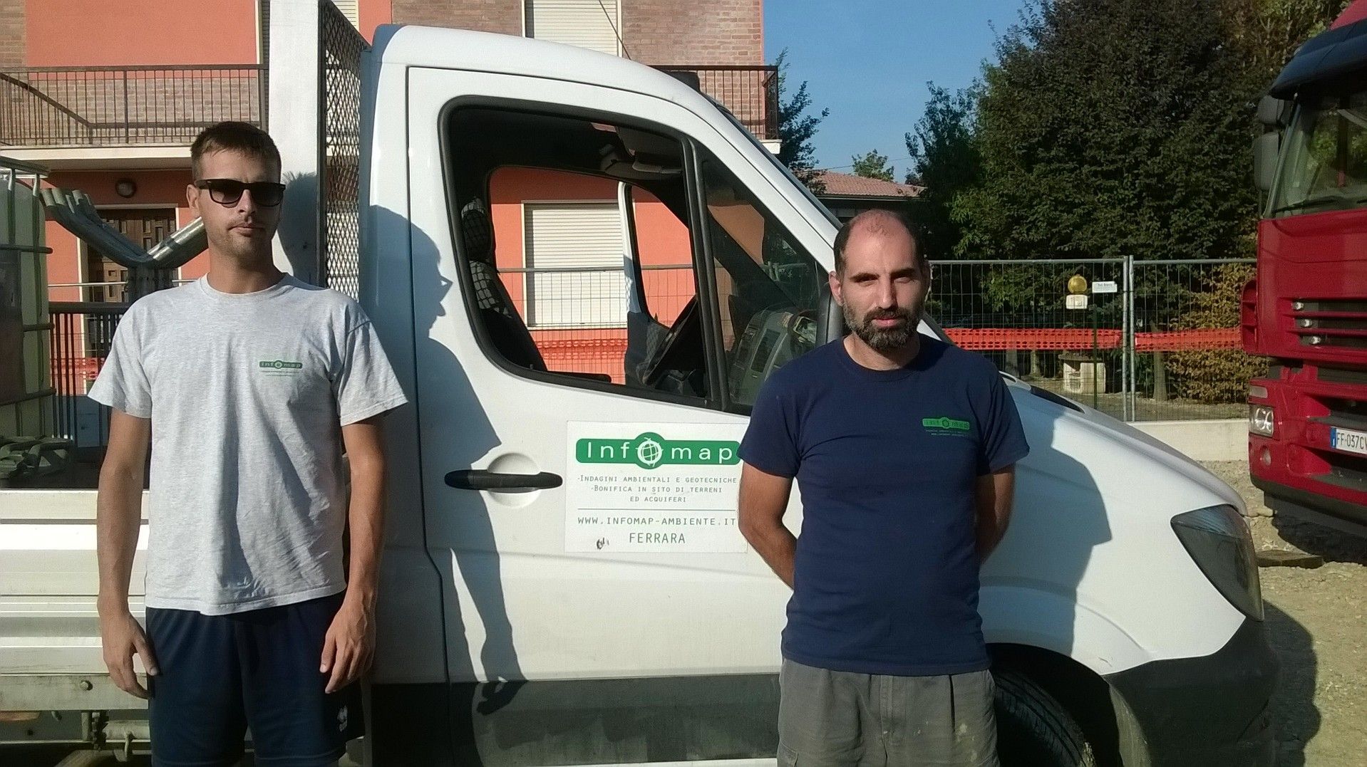 Due uomini in piedi accanto a un furgone bianco davanti a un edificio, uno con gli occhiali da sole, l'altro con le mani dietro la schiena.