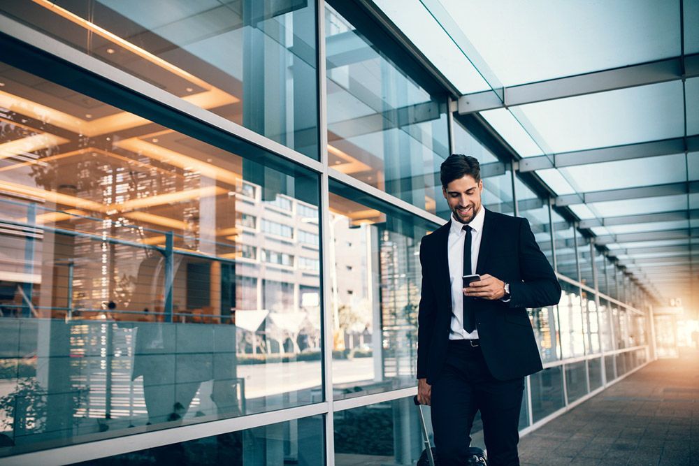 Man in a suit walking, looking at phone in a modern building with glass windows, suitcase in hand.
