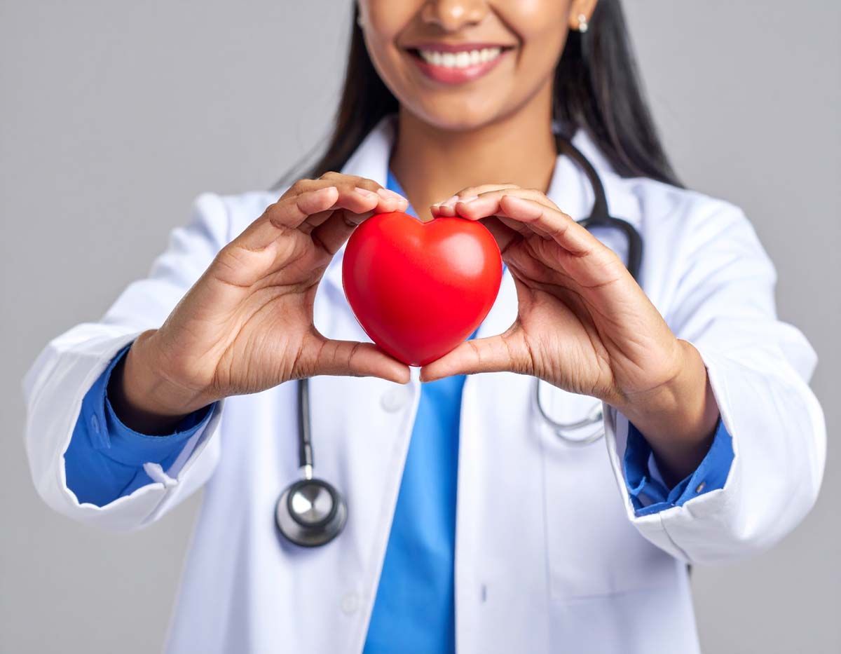 Doctor holding a red heart, smiling. Wearing a white coat and stethoscope.