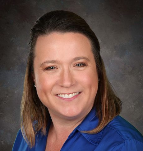 Woman with brown hair smiling, wearing a blue shirt, against a dark background.