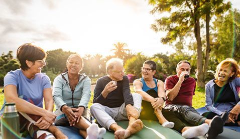 Group of people smiling and talking, sitting on mats outdoors in sunny park.