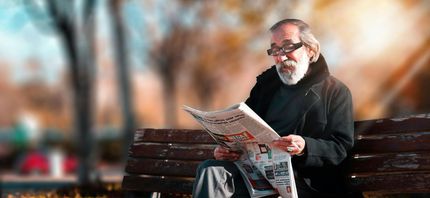 An older person wearing glasses reads a newspaper while sitting on a bench in a park.