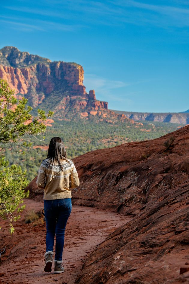 Woman hikes on a red dirt trail towards red rock formations under a blue sky.