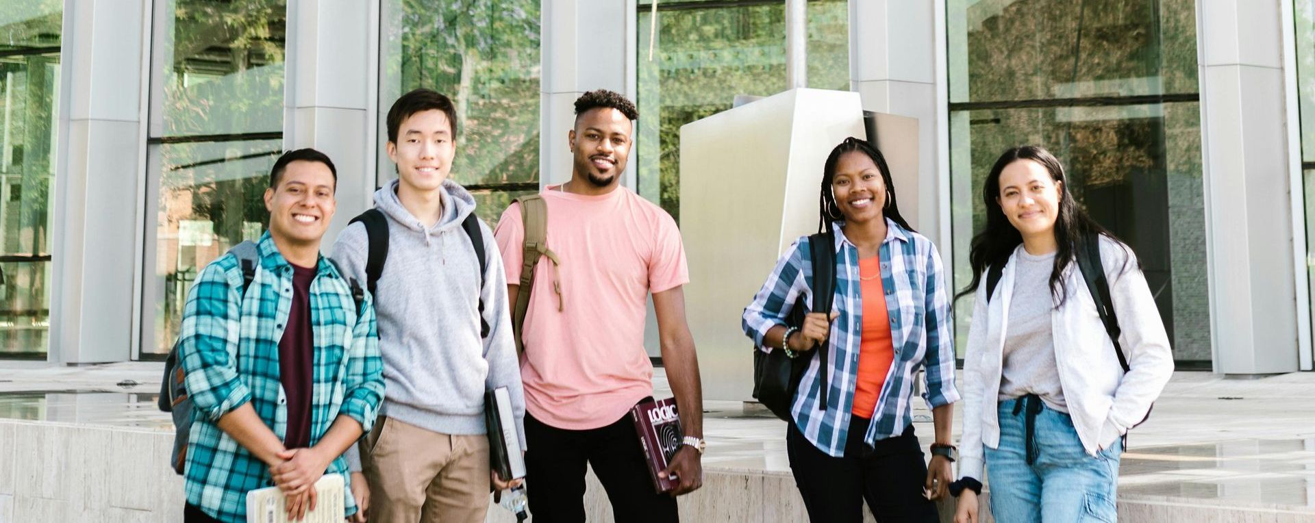 Five students standing outside a building, smiling at the camera. They wear casual clothes and backpacks.