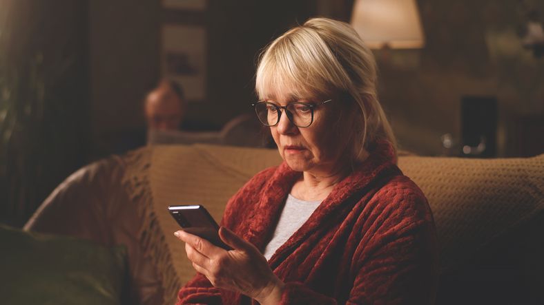 Woman with glasses looks intently at a phone screen while sitting on a couch, indoors.