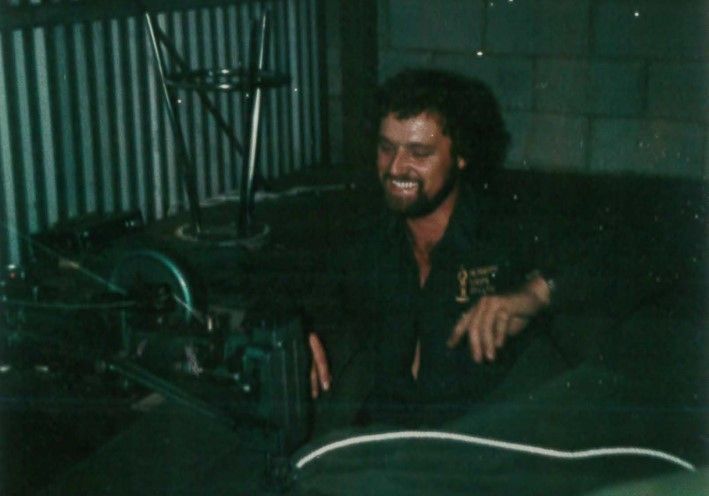 Man with a beard smiles while sitting near a large machine in a dimly lit workshop.