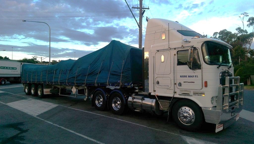 A large white semi-truck hauling a blue tarp-covered trailer on a road with a dusk sky background.