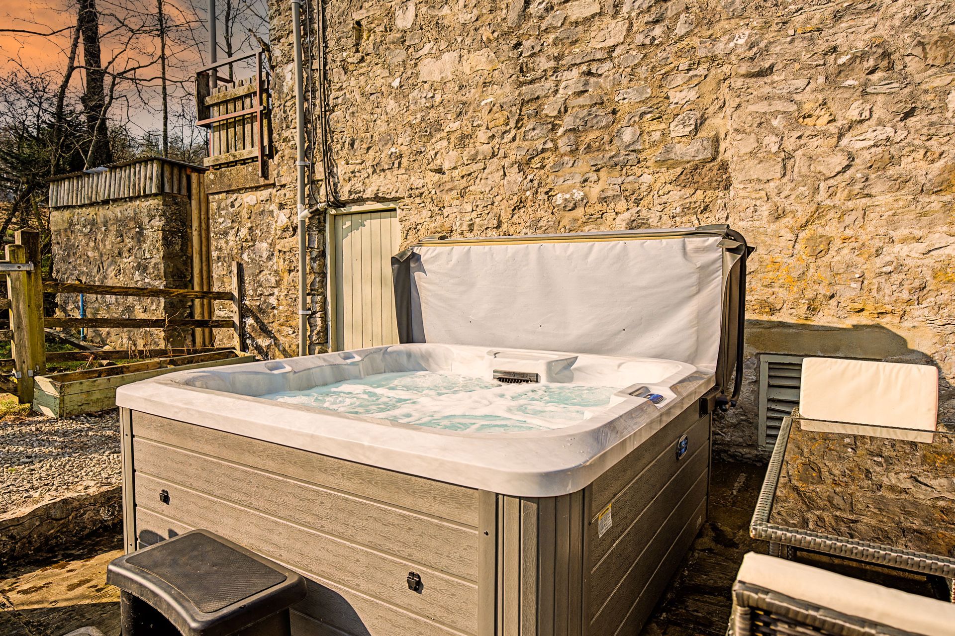Outdoor hot tub beside a stone wall and gate, with patio chairs and a bench in a sunny yard