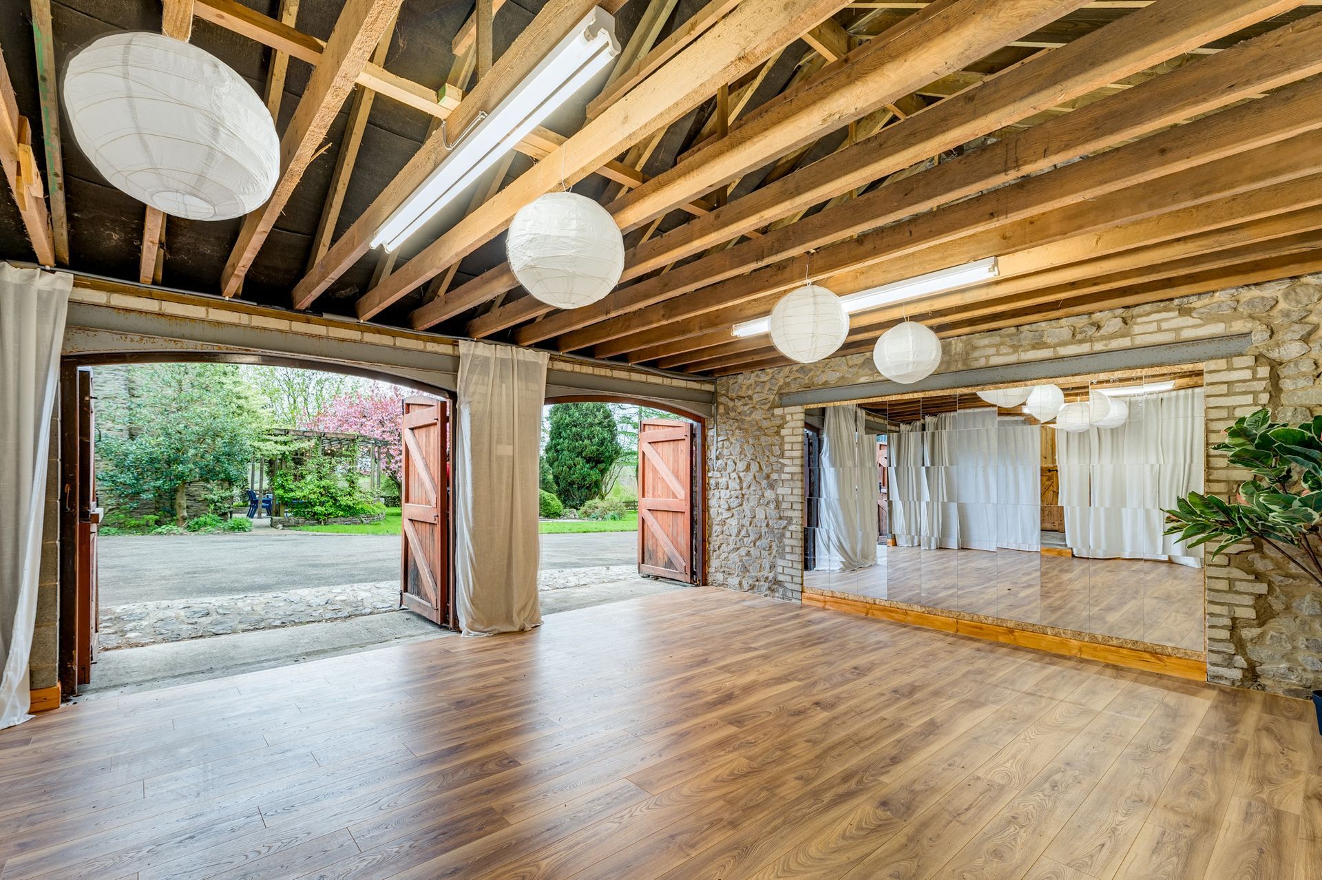 Spacious yoga studio with exposed beams, wood floor, hanging paper lights, and open doors to a courtyard.