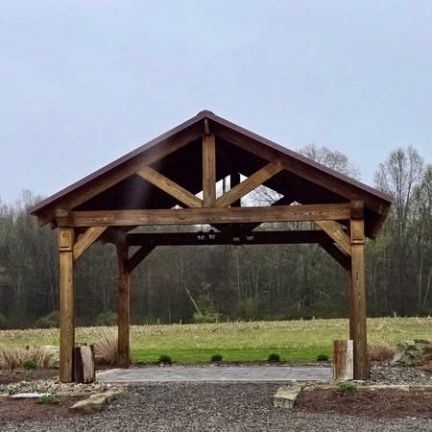 A wooden pavilion with a peaked roof stands on a stone patio overlooking a field and forest under a cloudy sky.