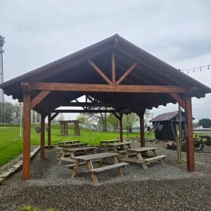 A wooden pavilion with picnic tables sits on a gravel patio in a grassy park setting under a cloudy sky.