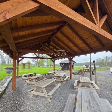 A covered wooden pavilion with multiple picnic tables on a gravel ground, overlooking a rural landscape with a shed.
