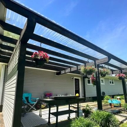 A black wooden pergola with a clear roof attached to a gray house, featuring hanging flower baskets and a patio grill.