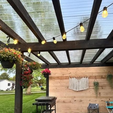 A pergola with string lights, hanging flower baskets, and a wooden wall decorated with macrame on a patio.