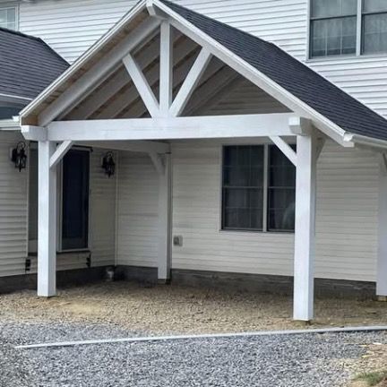 A white, wooden, gabled-roof carport structure attached to the side of a white-sided house with a gray gravel driveway.