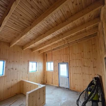 Interior of a room under construction with pine paneling on the walls and vaulted ceiling, concrete floor, and a doorway.