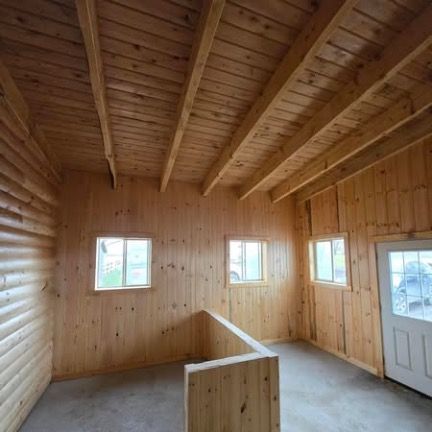 An interior view of a room with unfinished wooden walls, a beamed ceiling, two small windows, and a white entrance door.