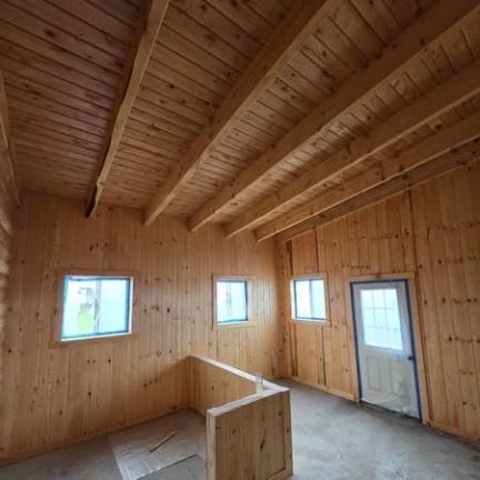 Interior of a rustic room with wood-paneled walls and ceiling, two windows, a white door, and a low partial partition wall.