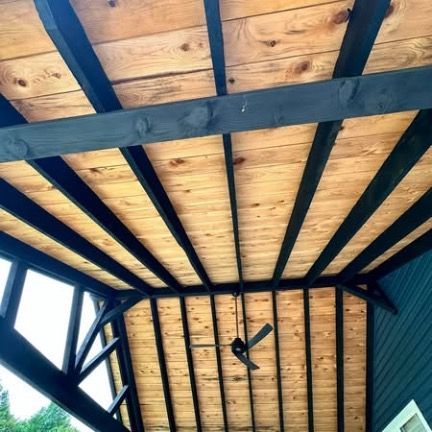 An outdoor porch ceiling featuring natural wood planks framed by black painted wooden beams, with a ceiling fan overhead.