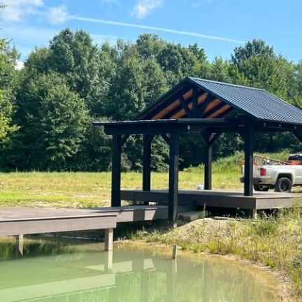 A wooden dock with a black-roofed shelter sits on a pond, with a pickup truck parked nearby against a backdrop of trees.
