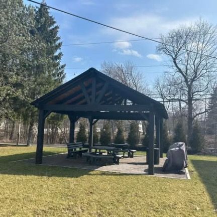 A dark wooden gazebo with outdoor seating sits on a stone patio in a grassy yard, with bare trees and evergreens nearby.