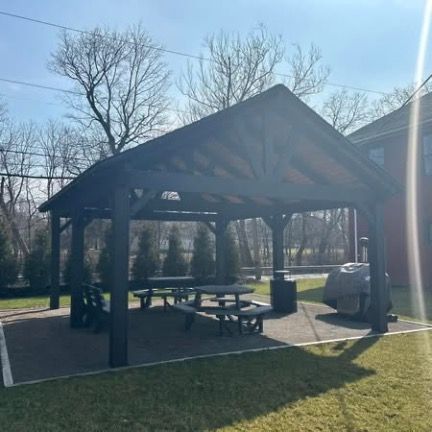 A dark wooden pavilion stands on a stone patio in a grassy yard, featuring picnic tables and a grill under the roof.
