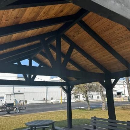 An outdoor pavilion with a black timber frame and natural wood-paneled ceiling, featuring a picnic table and bench.