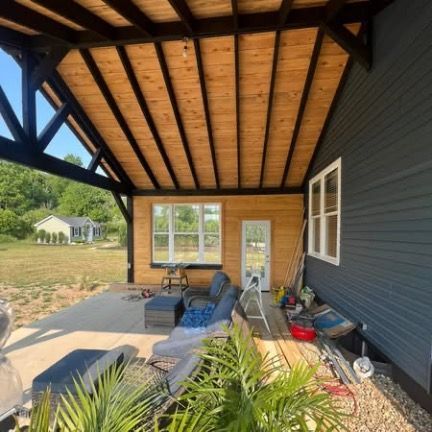 A modern outdoor covered patio with dark beams, wood ceiling, light siding, and furniture on a concrete floor.