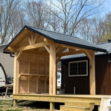 A wooden open-air timber frame pavilion attached to a dark brown home, featuring a dark metal gable roof and steps.