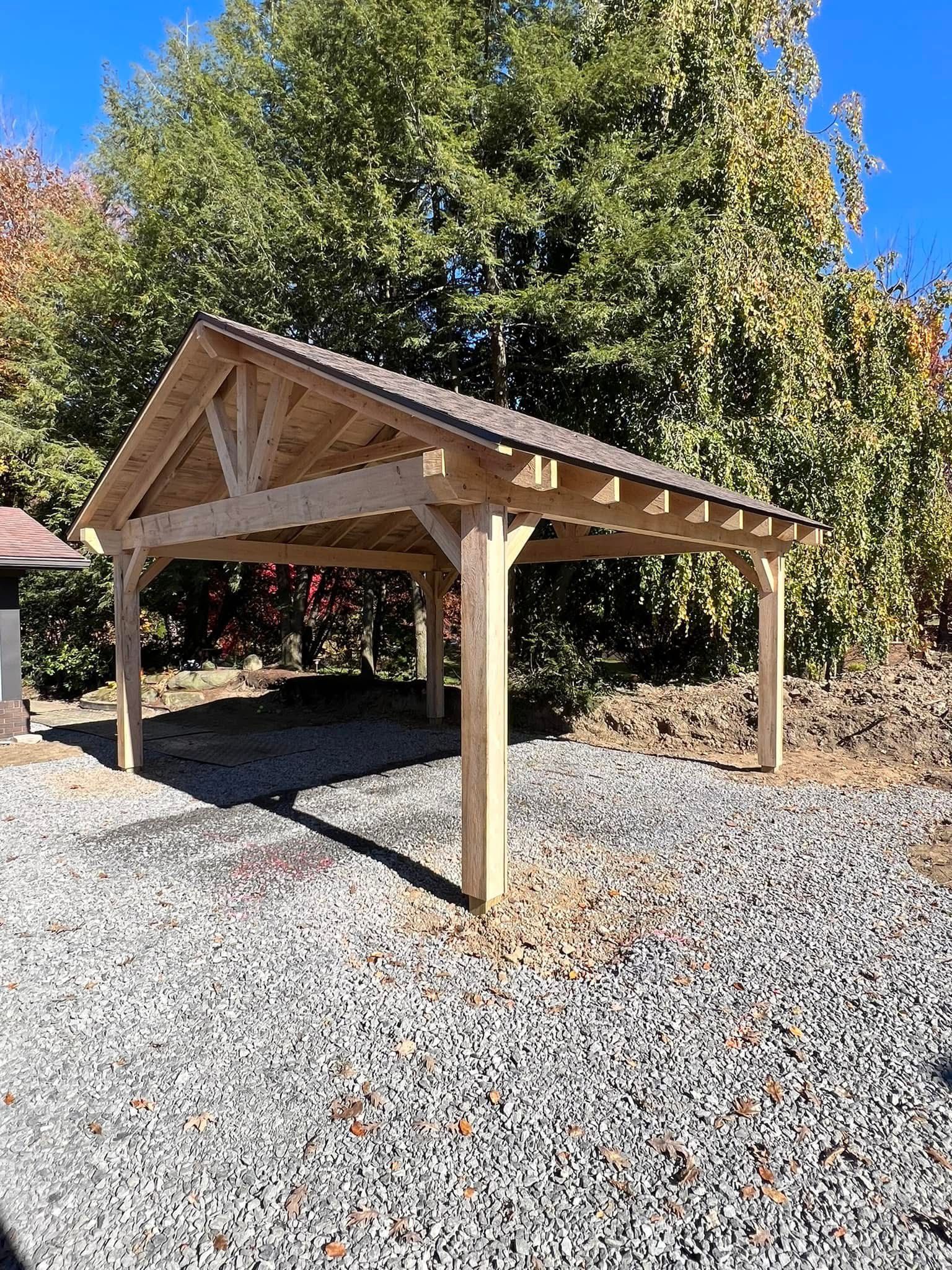 A wooden open-air pavilion with a pitched roof stands over a gravel area in front of a tree-filled background.