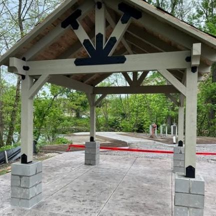A beige wooden pavilion with black decorative metal brackets stands on a gray concrete patio in a wooded area.