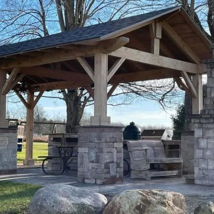 A wooden outdoor pavilion with a shingled roof, stone pillars, a stone fireplace, and rustic wooden seating.