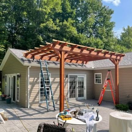 A wooden pergola under construction attached to a tan-sided house with a patio, two ladders, and a table with tools.