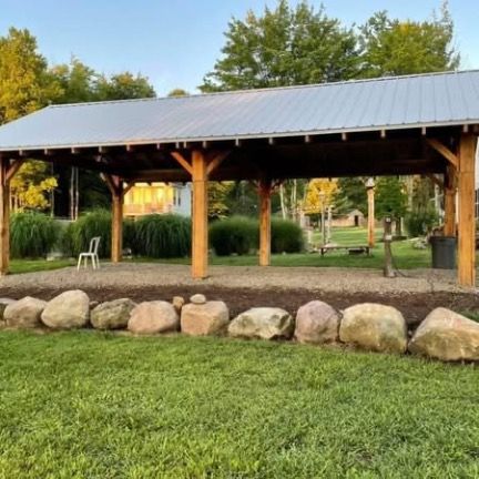 A wooden pavilion with a metal roof overlooks a gravel area lined with large boulders in a grassy yard.