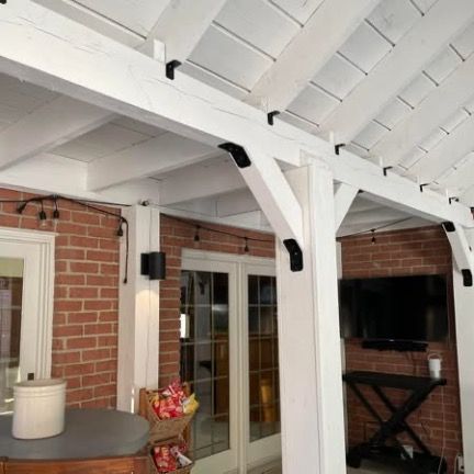 A white wooden patio structure with black decorative brackets, set against a red brick wall with a mounted television.