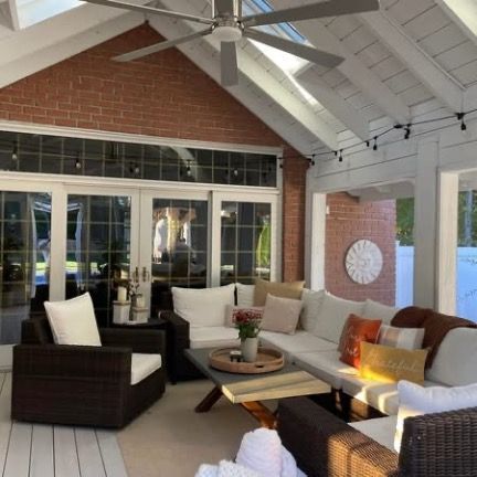 A cozy covered patio with dark wicker furniture, white cushions, a wooden coffee table, and exposed brick walls.