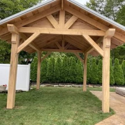 A sturdy, light-colored wooden pavilion stands in a grassy yard, featuring a gabled roof and a white privacy fence behind.